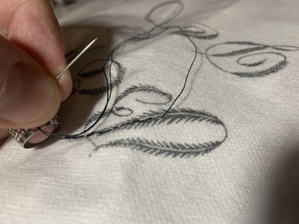 A hand holding a #10 sharp needle over a piece of embroidery